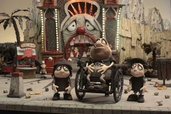 Still from the film Memoir of a Snail by Adam Elliot. Shows three characters, a father in a wheelchair and his two children outside Luna Park amusement park.